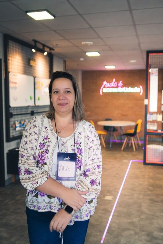 Retrato de Ale Miliatti em um espaço de trabalho com letreiro neon 'tudo é acessibilidade' ao fundo.